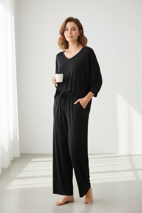 A black V-neck sleeveless top with palazzo style pants, hanging on a wooden hanger against a white background. A hand is adjusting the top by the sleeve, showing the fit and style of the garment.