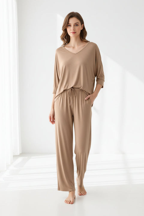 A beige V-neck sleeveless pajama suit with palazzo pants, displayed on a hanger and a person's hand adjusting the fabric.